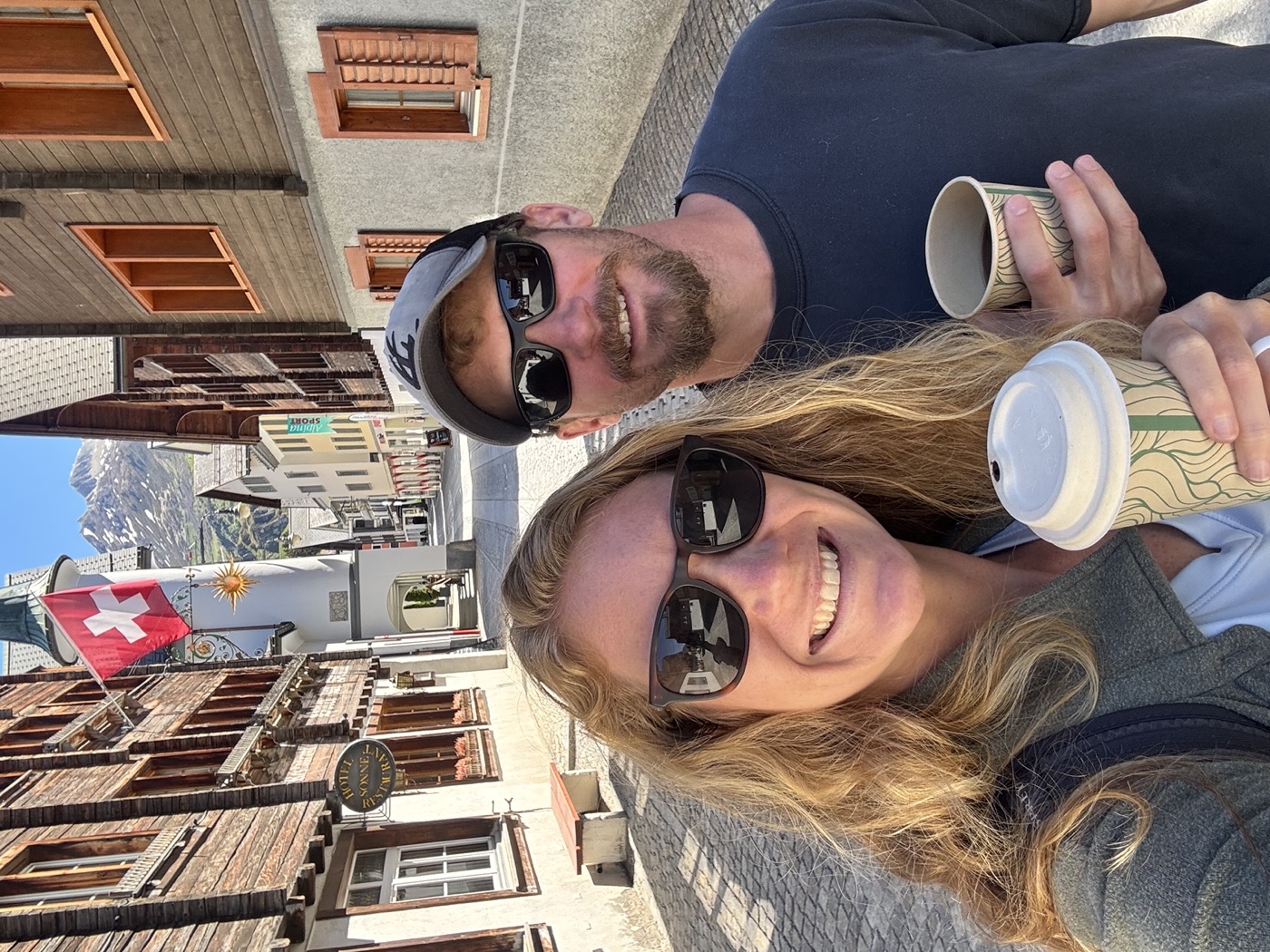 Jared and Anna smiling with coffees on a street in a Swiss mountain village