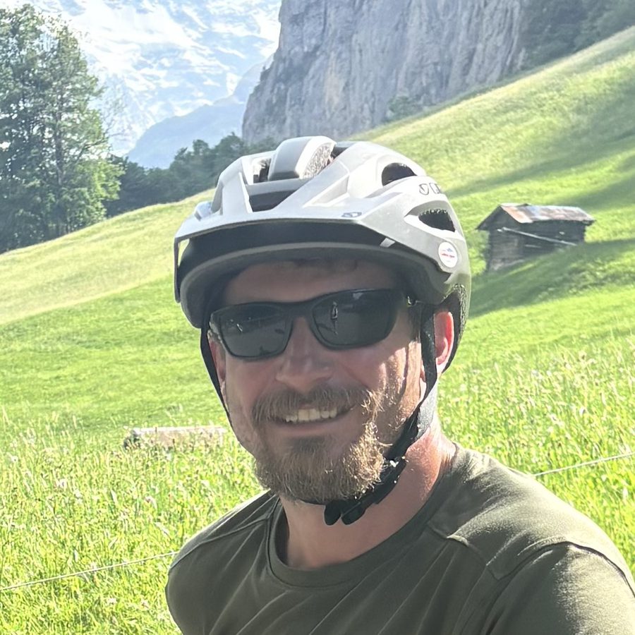 Close-up of Jared in a bike helmet and sunglasses, smiling, with green Swiss mountainside behind him